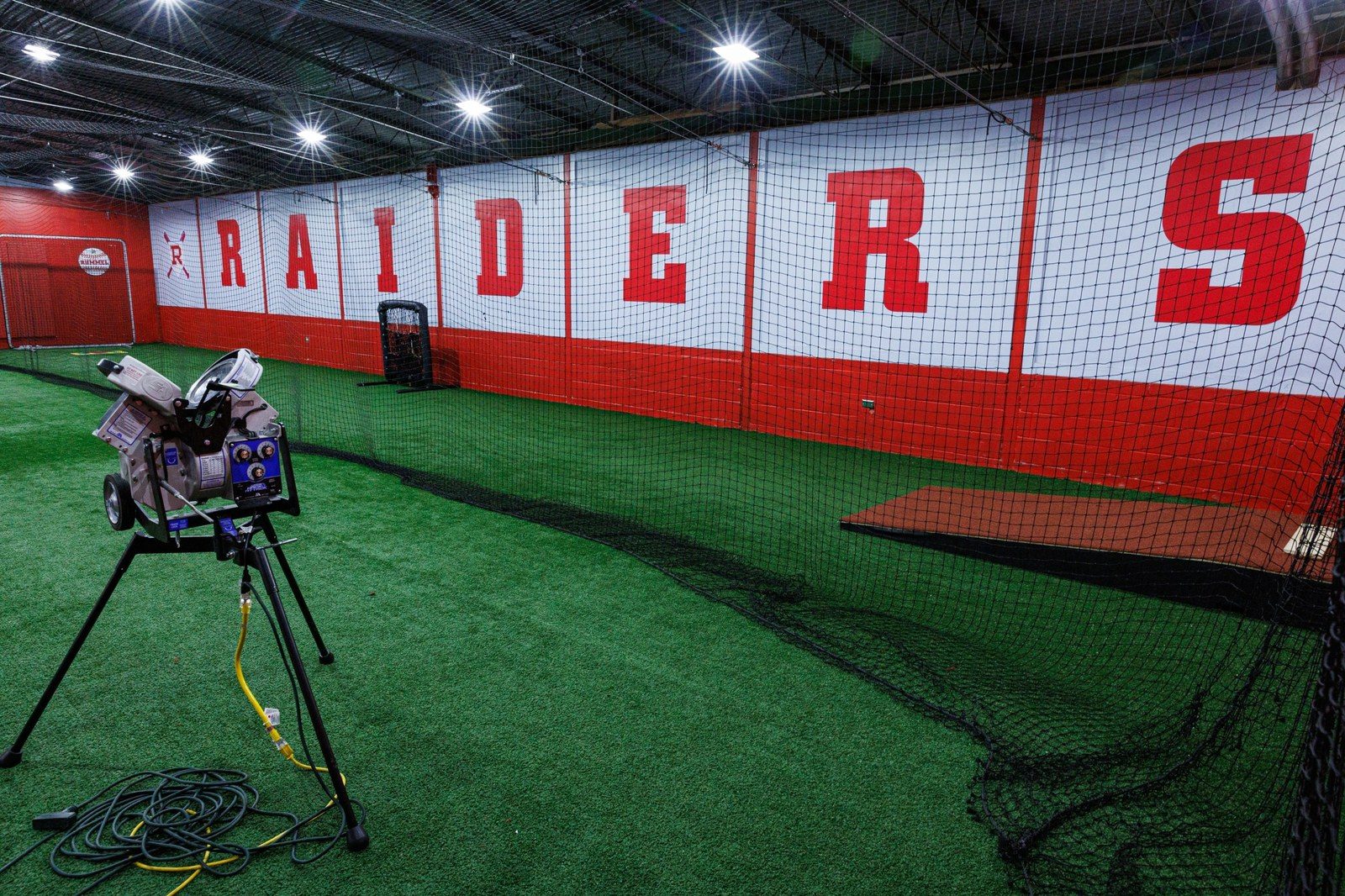 The indoor batting cage: turf, netting, a pitching machine, and RAIDERS across the wall