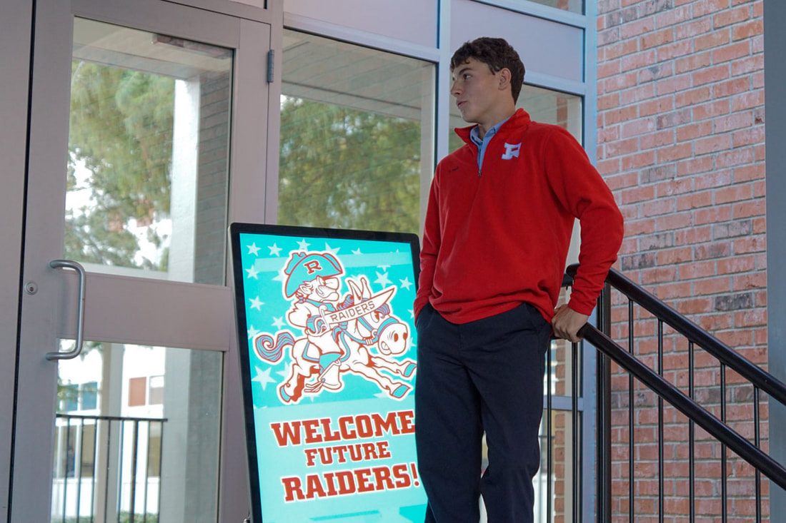 A student at the front doors of the school beside a Welcome Future Raiders sign