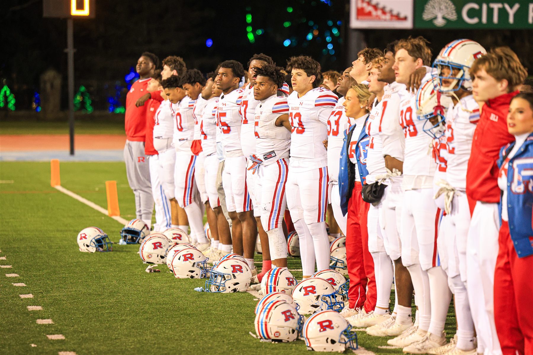 The football team lined up along the sideline at night, helmets on the turf