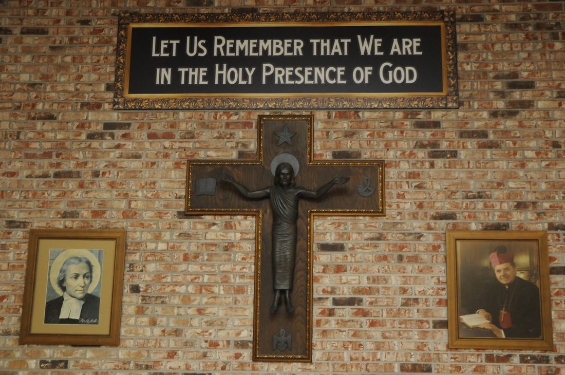 The Raider Room cross, a bronze Risen Christ on a wooden cross between portraits of Archbishop Rummel and St. La Salle, under the words Let us remember that we are in the holy presence of God