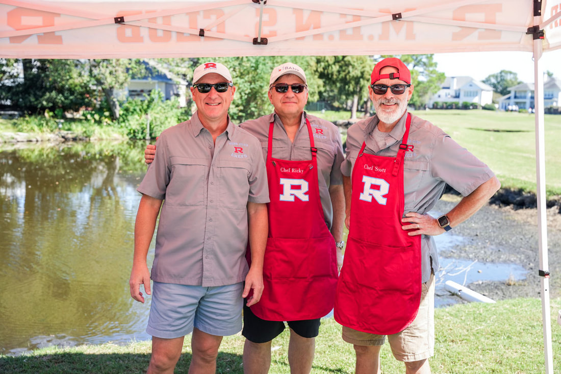 Three alumni cooks in red R aprons grinning under the tent by the water