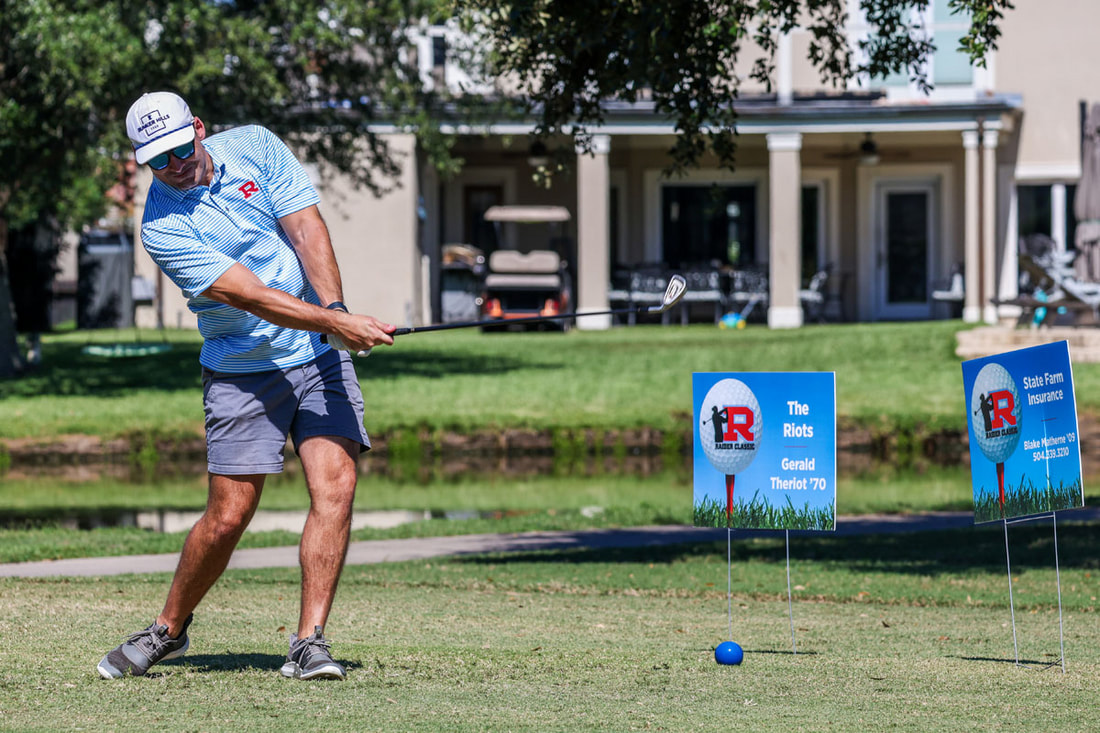 A Raider in a blue R polo mid swing at the tee, the R sponsor signs behind him