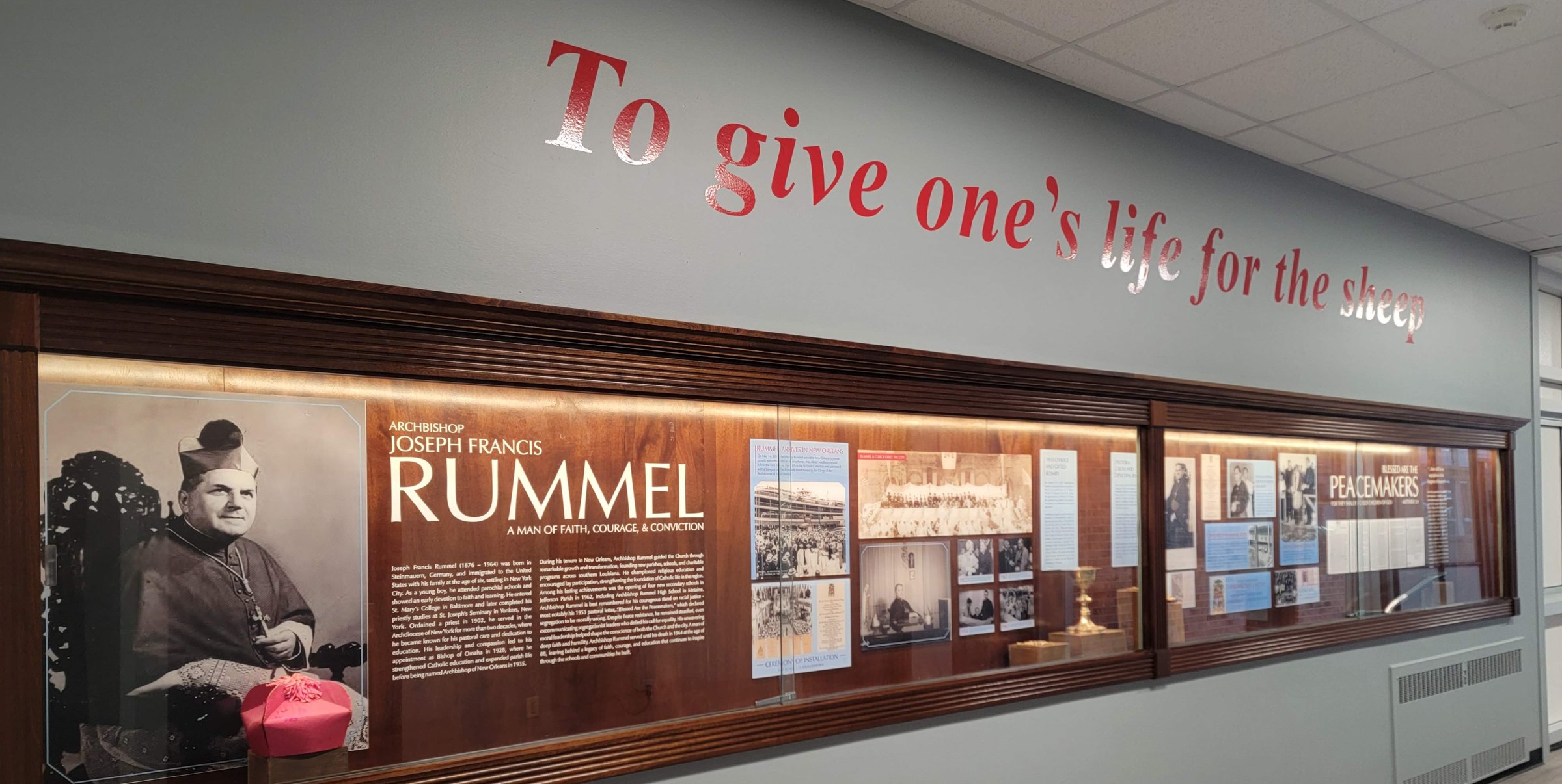 The Archbishop Rummel display case in the hallway, with the words To give one's life for the sheep on the wall above it