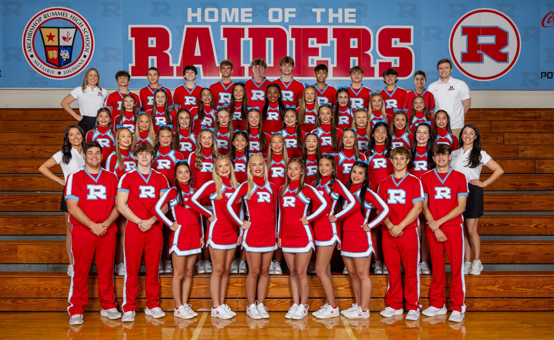 The 2026 Raider cheer squad on the gym floor in red, standing in rows in front of the HOME OF THE RAIDERS wall.