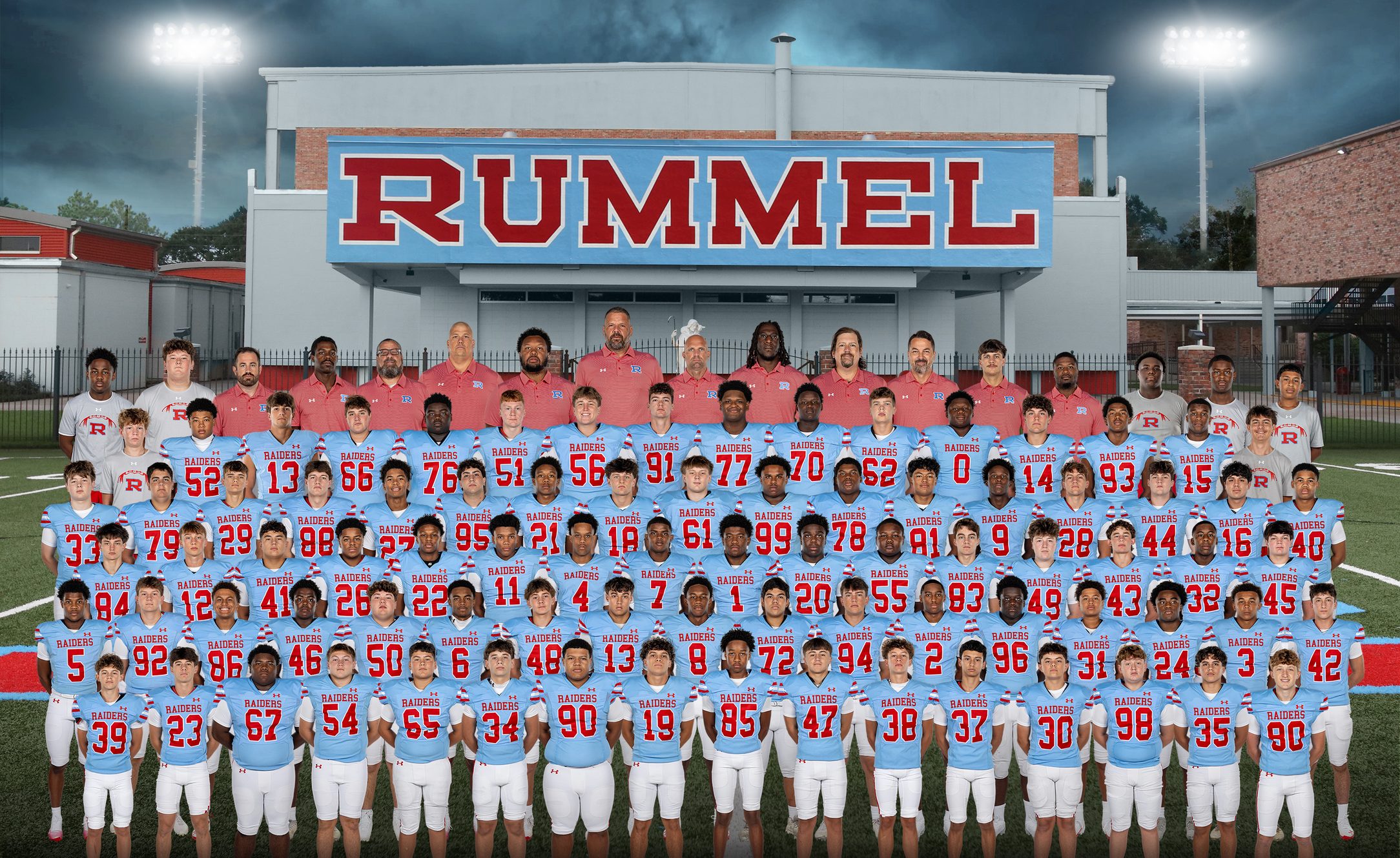 The 2026 Raider football team on the field in Columbia blue, the coaching staff standing behind them and the RUMMEL press box above.